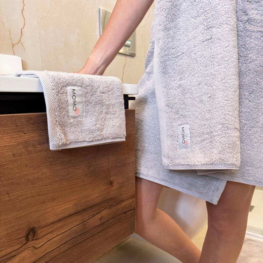 Conjunto de toallas Momo de algodón exhibido en estante de baño de madera con persona sosteniendo toallas beis dobladas