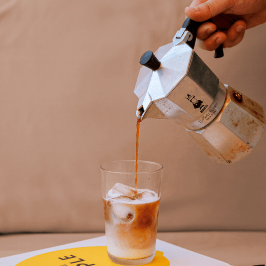 Moka caliente siendo vertida en una taza con hielo sobre fondo beige.