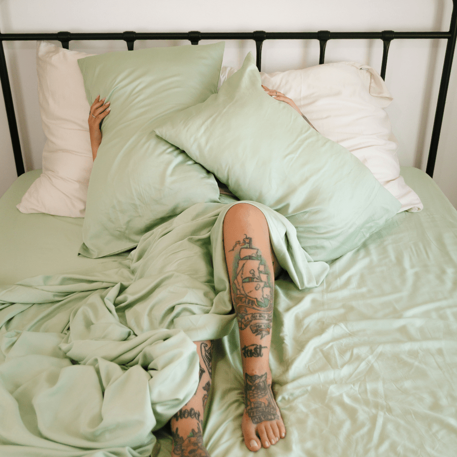Person lying in bed wearing green and white bamboo bedding set with visible tattoos on legs