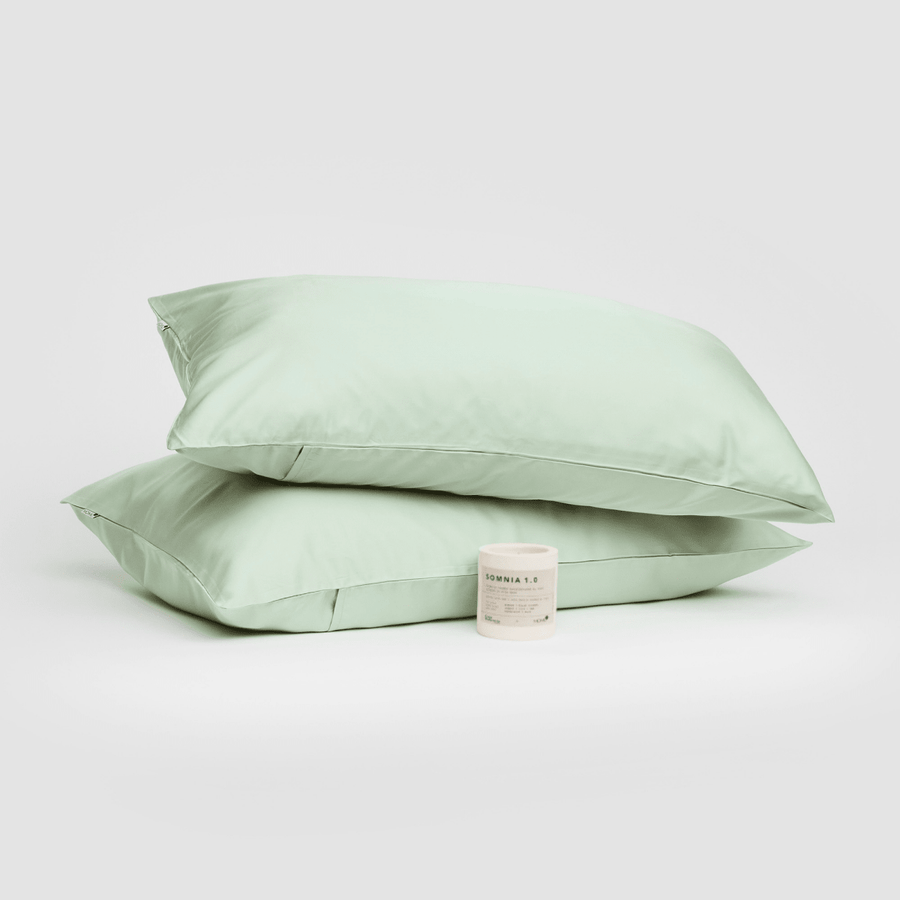 Pair of sage green cotton pillowcases stacked with Somnia 1.0 sleep aid candle on white background