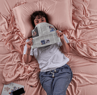 Mujer acostada en sábana de cama de bambú rosa leyendo periódico, vista desde arriba