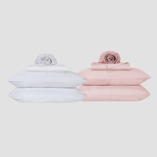 Juego de sábanas blanco y rosa apilado con almohadas y edredón enrollado sobre fondo neutro