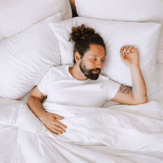 Hombre durmiendo tranquilamente en cama con sábanas blancas, rodeado de ropa de cama minimalista y cómoda que transmite descanso profundo.