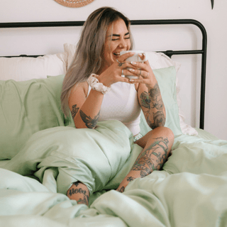 Mujer en la cama con camiseta blanca y juego de sábanas de bambú verde salvia, mostrando tejido suave y comodidad